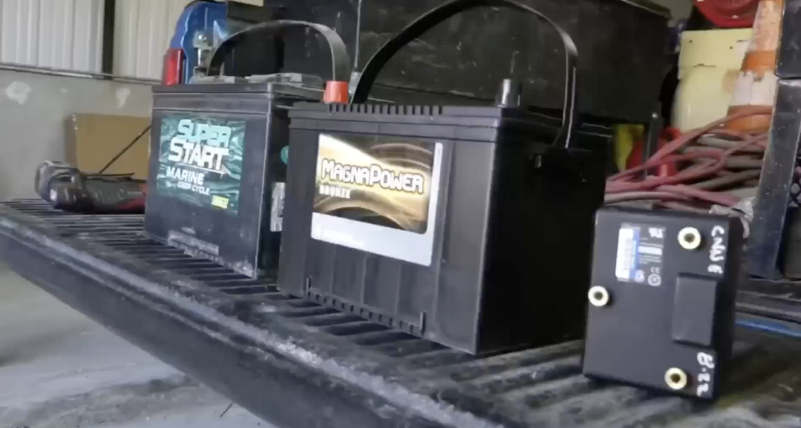 Two large black vehicle batteries, one labeled "Super Start Marine Deep Cycle" and the other "MagnaPower Bronze," sit side-by-side on the ribbed bed of a truck. To the right, a smaller black rectangular electronic component with brass terminals is also visible.