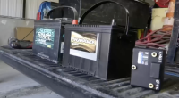 Two large black vehicle batteries, one labeled "Super Start Marine Deep Cycle" and the other "MagnaPower Bronze," sit side-by-side on the ribbed bed of a truck. To the right, a smaller black rectangular electronic component with brass terminals is also visible.
