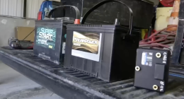 Two large black vehicle batteries, one labeled "Super Start Marine Deep Cycle" and the other "MagnaPower Bronze," sit side-by-side on the ribbed bed of a truck. To the right, a smaller black rectangular electronic component with brass terminals is also visible.