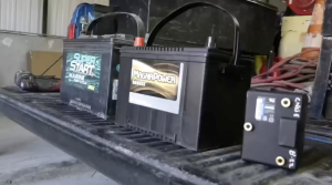 Two large black vehicle batteries, one labeled "Super Start Marine Deep Cycle" and the other "MagnaPower Bronze," sit side-by-side on the ribbed bed of a truck. To the right, a smaller black rectangular electronic component with brass terminals is also visible.