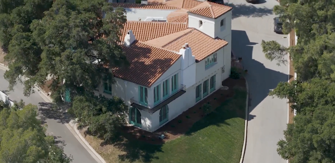 An aerial, high-angle shot of the William S Hart Museum shows a large, two-story Mediterranean-style villa with white stucco walls and a terracotta tile roof. The house features distinct teal-colored window frames and doors, including arched doorways on the ground floor and a small Juliet balcony on the second level. Two white chimneys rise from the roofline, and a square tower-like structure sits at one corner. The property is nestled between two paved roads that curve around its perimeter, surrounded by lush green trees and a small, manicured lawn.