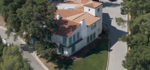 An aerial, high-angle shot of the William S Hart Museum shows a large, two-story Mediterranean-style villa with white stucco walls and a terracotta tile roof. The house features distinct teal-colored window frames and doors, including arched doorways on the ground floor and a small Juliet balcony on the second level. Two white chimneys rise from the roofline, and a square tower-like structure sits at one corner. The property is nestled between two paved roads that curve around its perimeter, surrounded by lush green trees and a small, manicured lawn.