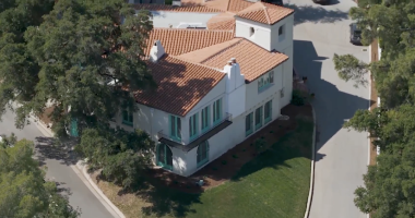 An aerial, high-angle shot of the William S Hart Museum shows a large, two-story Mediterranean-style villa with white stucco walls and a terracotta tile roof. The house features distinct teal-colored window frames and doors, including arched doorways on the ground floor and a small Juliet balcony on the second level. Two white chimneys rise from the roofline, and a square tower-like structure sits at one corner. The property is nestled between two paved roads that curve around its perimeter, surrounded by lush green trees and a small, manicured lawn.
