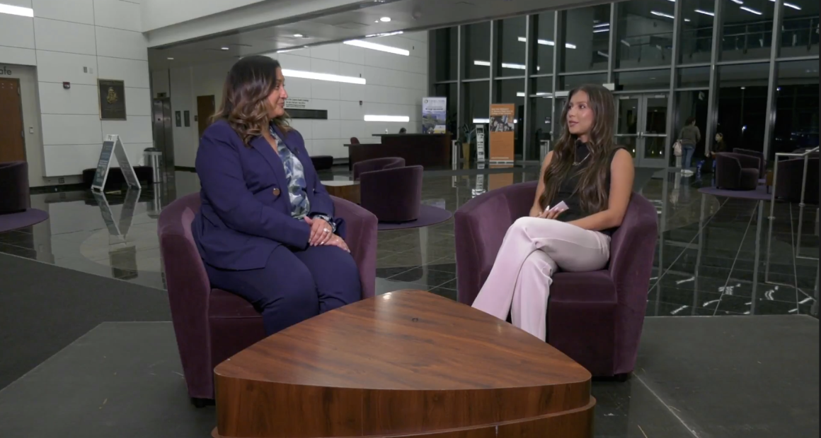Two women sit in purple armchairs facing each other in a modern indoor lobby, engaged in an interview across a wooden coffee table, with glass walls and a reception desk visible in the background.