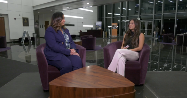 Two women sit in purple armchairs facing each other in a modern indoor lobby, engaged in an interview across a wooden coffee table, with glass walls and a reception desk visible in the background.