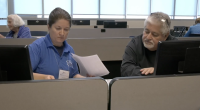 A man in a black sweater receives help with his taxes from a VITA representative in a blue shirt, as they review documents together at a computer workstation in a classroom setting.