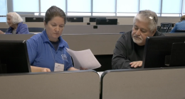 A man in a black sweater receives help with his taxes from a VITA representative in a blue shirt, as they review documents together at a computer workstation in a classroom setting.