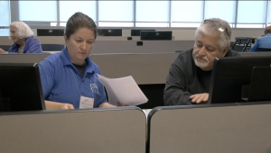 A man in a black sweater receives help with his taxes from a VITA representative in a blue shirt, as they review documents together at a computer workstation in a classroom setting.