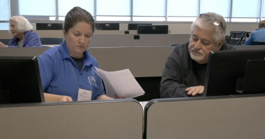 A man in a black sweater receives help with his taxes from a VITA representative in a blue shirt, as they review documents together at a computer workstation in a classroom setting.