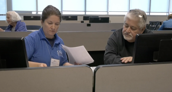 A man in a black sweater receives help with his taxes from a VITA representative in a blue shirt, as they review documents together at a computer workstation in a classroom setting.