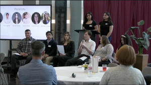 A wide shot of an indoor panel discussion held in a modern room with a large screen and a maroon curtain backdrop. Five panelists sit in a row of chairs facing an audience. From left to right: a man in a plaid shirt, a man in a dark quarter-zip, a woman in a black blazer holding a paper, a man in a white button-down speaking into a microphone, and a woman in a patterned blouse. Behind them, two women in black t-shirts stand near a podium.
