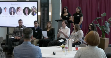 A wide shot of an indoor panel discussion held in a modern room with a large screen and a maroon curtain backdrop. Five panelists sit in a row of chairs facing an audience. From left to right: a man in a plaid shirt, a man in a dark quarter-zip, a woman in a black blazer holding a paper, a man in a white button-down speaking into a microphone, and a woman in a patterned blouse. Behind them, two women in black t-shirts stand near a podium.