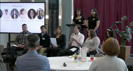 A wide shot of an indoor panel discussion held in a modern room with a large screen and a maroon curtain backdrop. Five panelists sit in a row of chairs facing an audience. From left to right: a man in a plaid shirt, a man in a dark quarter-zip, a woman in a black blazer holding a paper, a man in a white button-down speaking into a microphone, and a woman in a patterned blouse. Behind them, two women in black t-shirts stand near a podium.