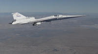 A sleek experimental NASA X-59 jet, marked with NASA branding, flies with its landing gear down over a vast desert landscape, with mountains faintly visible in the distance.