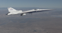 A sleek experimental NASA X-59 jet, marked with NASA branding, flies with its landing gear down over a vast desert landscape, with mountains faintly visible in the distance.