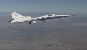 A sleek experimental NASA X-59 jet, marked with NASA branding, flies with its landing gear down over a vast desert landscape, with mountains faintly visible in the distance.