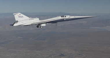A sleek experimental NASA X-59 jet, marked with NASA branding, flies with its landing gear down over a vast desert landscape, with mountains faintly visible in the distance.