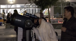 A person peers through a large telescope on a tripod during an evening outdoor event, while another person stands nearby watching. In the background, small groups gather under lights outside a building, suggesting a nighttime stargazing or community event.