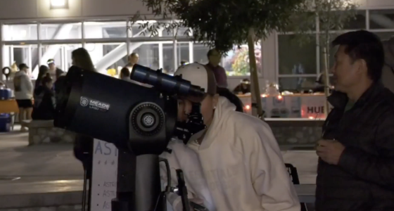 A person peers through a large telescope on a tripod during an evening outdoor event, while another person stands nearby watching. In the background, small groups gather under lights outside a building, suggesting a nighttime stargazing or community event.