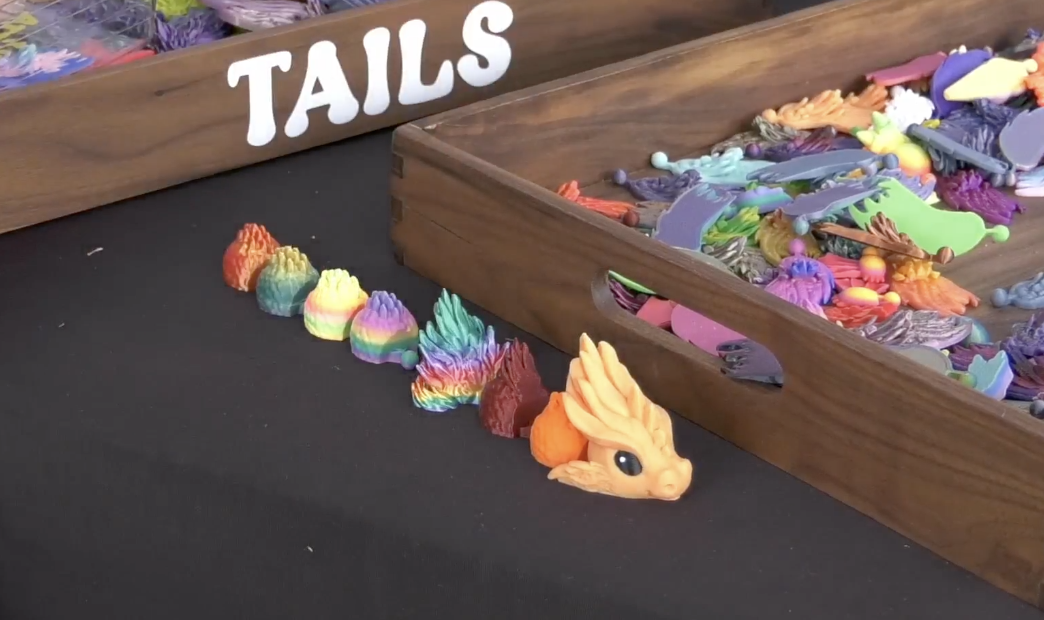 A high-angle, close-up shot of a black tabletop featuring 3D-printed dragon components. In the foreground, a small, orange dragon head with large black eyes sits at the end of a line of various colorful, textured segments that transition through a rainbow of colors including red, yellow, green, and blue.