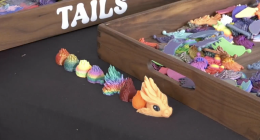A high-angle, close-up shot of a black tabletop featuring 3D-printed dragon components. In the foreground, a small, orange dragon head with large black eyes sits at the end of a line of various colorful, textured segments that transition through a rainbow of colors including red, yellow, green, and blue.