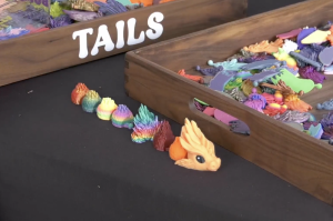 A high-angle, close-up shot of a black tabletop featuring 3D-printed dragon components. In the foreground, a small, orange dragon head with large black eyes sits at the end of a line of various colorful, textured segments that transition through a rainbow of colors including red, yellow, green, and blue.