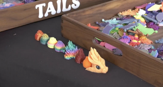 A high-angle, close-up shot of a black tabletop featuring 3D-printed dragon components. In the foreground, a small, orange dragon head with large black eyes sits at the end of a line of various colorful, textured segments that transition through a rainbow of colors including red, yellow, green, and blue.