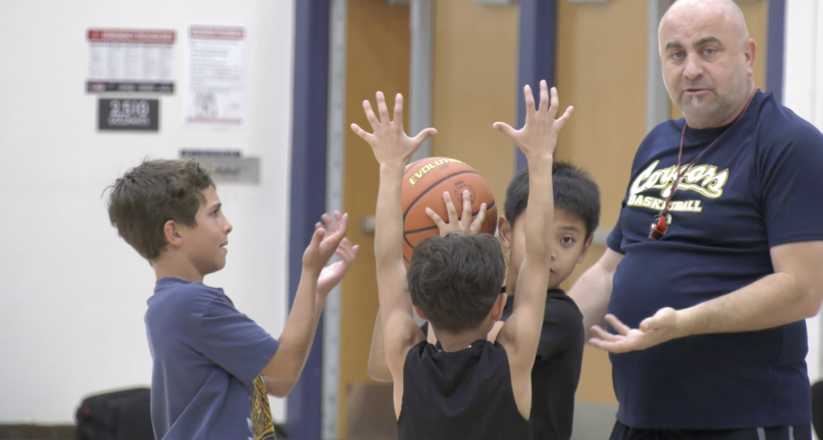 A basketball coach stands in a gym instructing three young boys during a drill. Two boys hold a basketball overhead with their arms raised while another boy reaches toward the ball, as the coach gestures with his hands and watches closely.