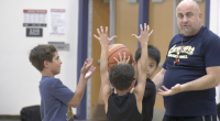 A basketball coach stands in a gym instructing three young boys during a drill. Two boys hold a basketball overhead with their arms raised while another boy reaches toward the ball, as the coach gestures with his hands and watches closely.