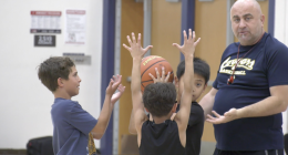 A basketball coach stands in a gym instructing three young boys during a drill. Two boys hold a basketball overhead with their arms raised while another boy reaches toward the ball, as the coach gestures with his hands and watches closely.