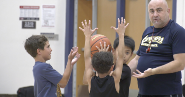 A basketball coach stands in a gym instructing three young boys during a drill. Two boys hold a basketball overhead with their arms raised while another boy reaches toward the ball, as the coach gestures with his hands and watches closely.