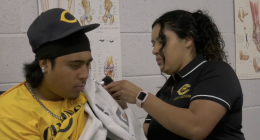 An athletic trainer wearing a black "Canyons Sports Medicine" polo shirt carefully treats a male athlete's shoulder. The trainer, with curly dark hair tied back and a pink smartwatch, uses a handheld therapeutic device over a white towel draped across the athlete's shoulder. The athlete is wearing a yellow "Canyons" shirt, a black baseball cap with a gold "C" logo, and a silver chain. They are in a sports medicine facility with anatomical posters of the hip and knee visible on the white cinderblock wall in the background.