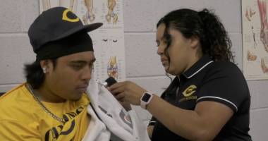 An athletic trainer wearing a black "Canyons Sports Medicine" polo shirt carefully treats a male athlete's shoulder. The trainer, with curly dark hair tied back and a pink smartwatch, uses a handheld therapeutic device over a white towel draped across the athlete's shoulder. The athlete is wearing a yellow "Canyons" shirt, a black baseball cap with a gold "C" logo, and a silver chain. They are in a sports medicine facility with anatomical posters of the hip and knee visible on the white cinderblock wall in the background.