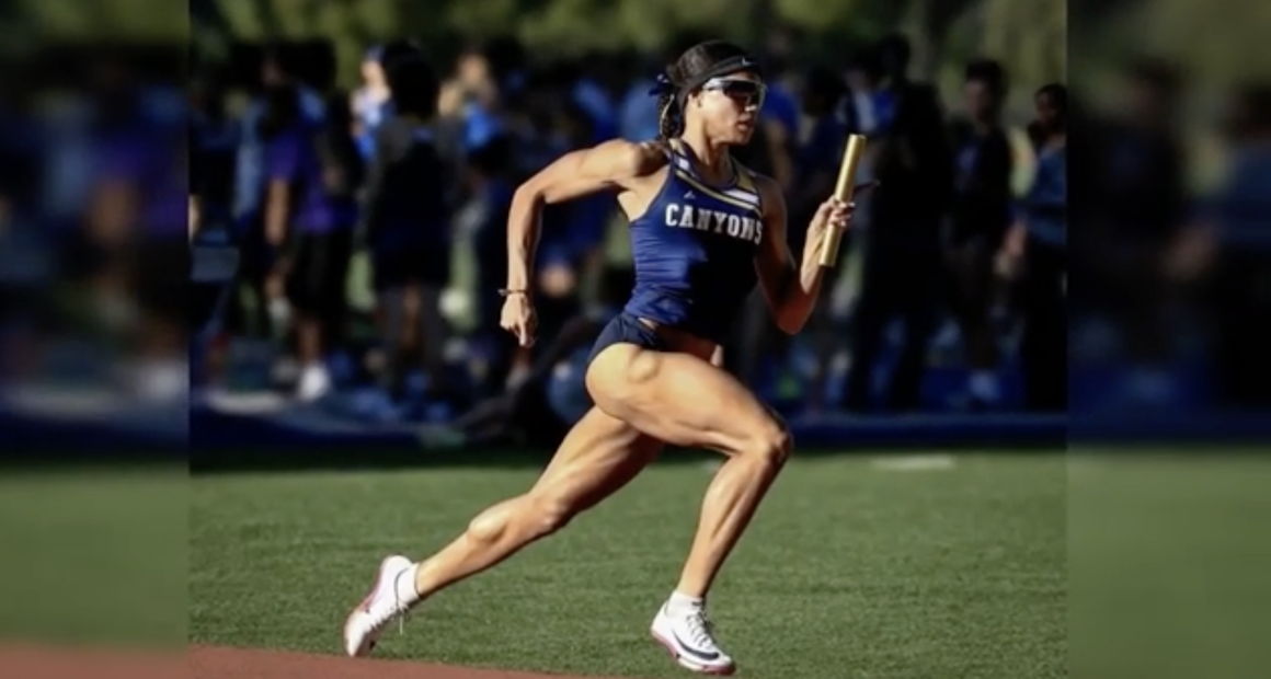 A female track athlete in a blue “Canyons” uniform sprints on a track while holding a relay baton. She is mid-stride, wearing sunglasses and a headband, with a focused expression, as a blurred crowd and other athletes stand in the background.