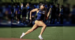 A female track athlete in a blue “Canyons” uniform sprints on a track while holding a relay baton. She is mid-stride, wearing sunglasses and a headband, with a focused expression, as a blurred crowd and other athletes stand in the background.