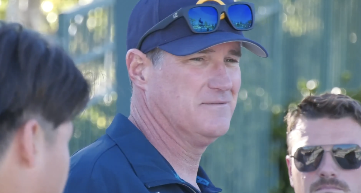 Coach Erik Matheson stands outdoors wearing a navy cap with sunglasses resting on the brim and a dark polo shirt, looking off to the side while speaking. Two other men are partially visible nearby, suggesting a casual conversation or coaching moment in a sunny, outdoor setting.