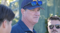 Coach Erik Matheson stands outdoors wearing a navy cap with sunglasses resting on the brim and a dark polo shirt, looking off to the side while speaking. Two other men are partially visible nearby, suggesting a casual conversation or coaching moment in a sunny, outdoor setting.