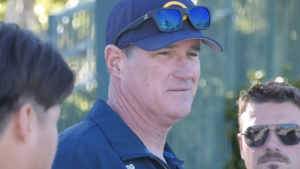Coach Erik Matheson stands outdoors wearing a navy cap with sunglasses resting on the brim and a dark polo shirt, looking off to the side while speaking. Two other men are partially visible nearby, suggesting a casual conversation or coaching moment in a sunny, outdoor setting.