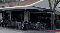 view of Nealie’s Skillet, a restaurant with a black wrap-around awning and a street address of 25858. The scene shows a patio dining area enclosed by a black metal fence, where several people are seated at small tables. To the left of the restaurant, a sign for PJ’s Barber Shop is visible in the window. The building has a tan, slanted roof and is situated next to a large tree on the right, under an overcast sky.