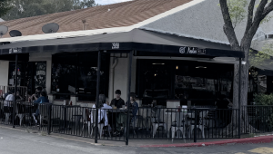 view of Nealie’s Skillet, a restaurant with a black wrap-around awning and a street address of 25858. The scene shows a patio dining area enclosed by a black metal fence, where several people are seated at small tables. To the left of the restaurant, a sign for PJ’s Barber Shop is visible in the window. The building has a tan, slanted roof and is situated next to a large tree on the right, under an overcast sky.