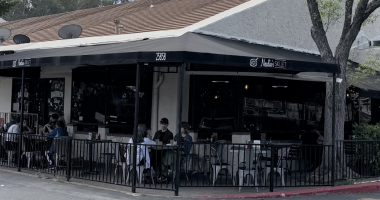 view of Nealie’s Skillet, a restaurant with a black wrap-around awning and a street address of 25858. The scene shows a patio dining area enclosed by a black metal fence, where several people are seated at small tables. To the left of the restaurant, a sign for PJ’s Barber Shop is visible in the window. The building has a tan, slanted roof and is situated next to a large tree on the right, under an overcast sky.