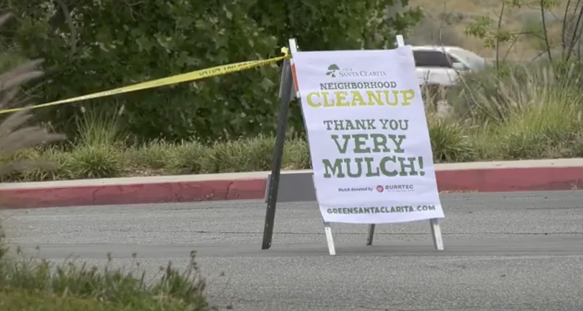 A roadside sign in Santa Clarita promotes a neighborhood cleanup event, reading “Neighborhood Cleanup — Thank You Very Mulch!” The sign stands on the edge of a street near greenery, with yellow caution tape and a parked vehicle visible in the background.