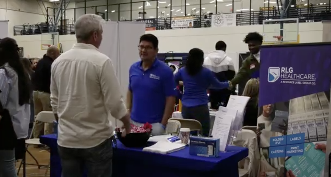 A representative wearing a blue polo shirt and glasses smiles while engaging with the visitor; next to him, a woman in a blue long-sleeved shirt is partially turned away.
