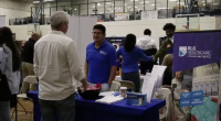 A representative wearing a blue polo shirt and glasses smiles while engaging with the visitor; next to him, a woman in a blue long-sleeved shirt is partially turned away.