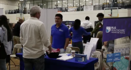 A representative wearing a blue polo shirt and glasses smiles while engaging with the visitor; next to him, a woman in a blue long-sleeved shirt is partially turned away.