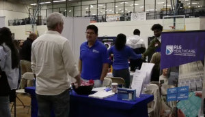 A representative wearing a blue polo shirt and glasses smiles while engaging with the visitor; next to him, a woman in a blue long-sleeved shirt is partially turned away.
