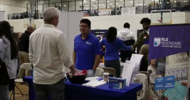 A representative wearing a blue polo shirt and glasses smiles while engaging with the visitor; next to him, a woman in a blue long-sleeved shirt is partially turned away.