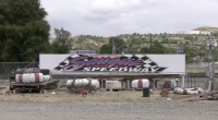 A large “Saugus Speedway” sign sits on display behind a row of worn red-and-white track barriers, with scattered debris and fencing in the foreground and rolling hills and a cloudy sky in the background.