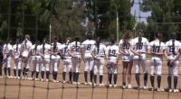 softball players in white uniforms stands shoulder to shoulder on the field during a pregame moment, seen through a protective net with trees and the outfield fence in the backgroun