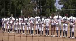 softball players in white uniforms stands shoulder to shoulder on the field during a pregame moment, seen through a protective net with trees and the outfield fence in the backgroun